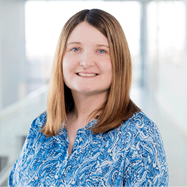 A woman wearing a bright blue shirt stands in front of a large window and smiles at the camera.